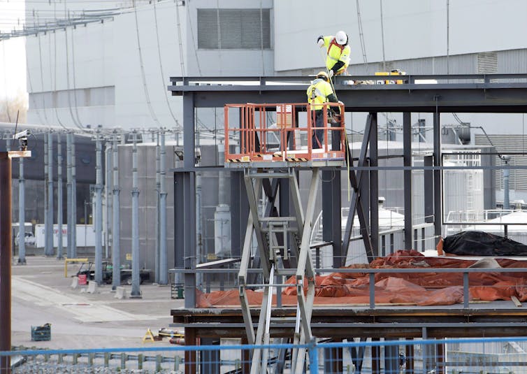 Two men seen working on the frame of a building against the backdrop of a large structure.