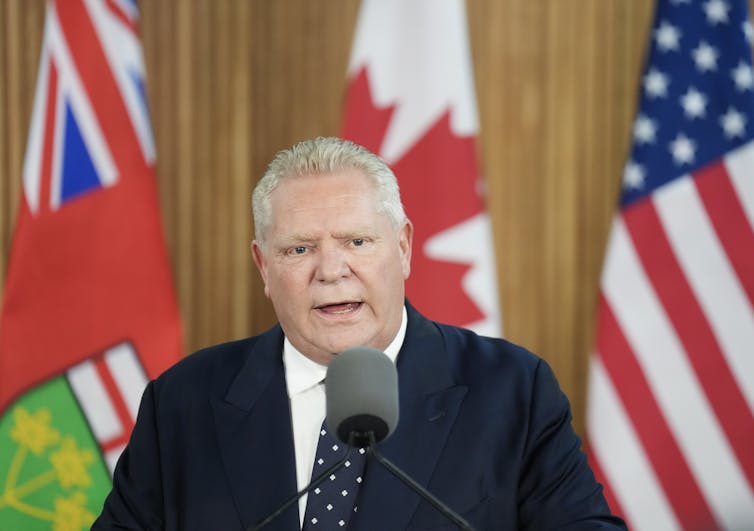 A rotund man with slicked back hair speaks into a microphone in front of Ontario, Canada and American flags.