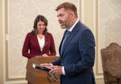 A man with reddish-hair and a beard holds a Bible while a dark-haired woman in a burgundy suit stands to his side.