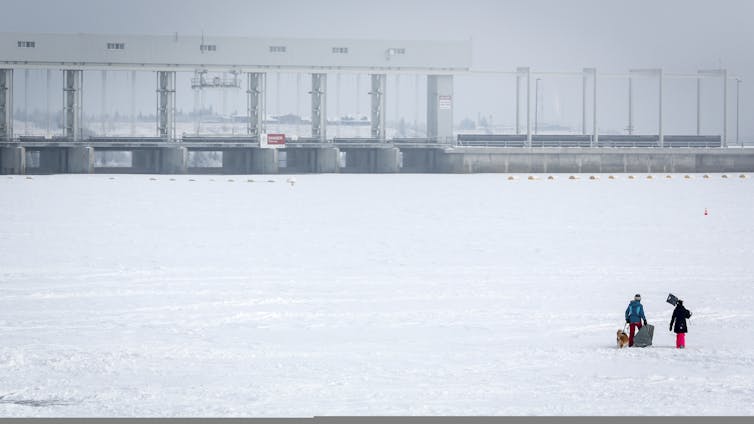 Two people clear ice on a frozen lake with a hydroelectric dam in the background.