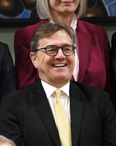 A man with dark hair and glasses smiles.