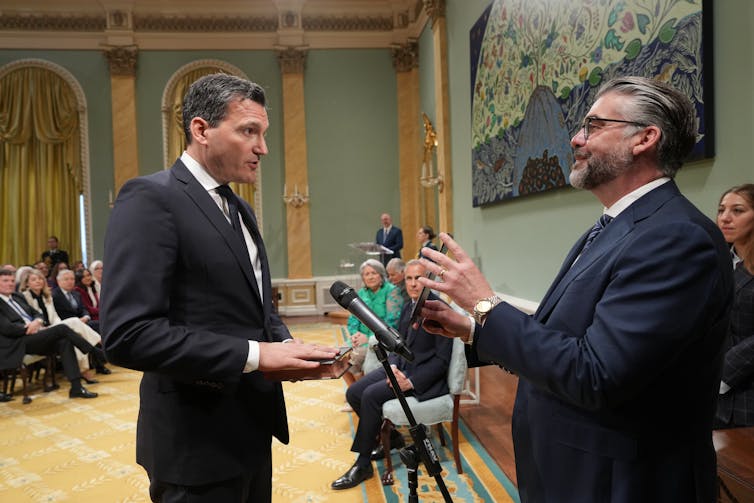 two men in suits stand facing each other in a formal room
