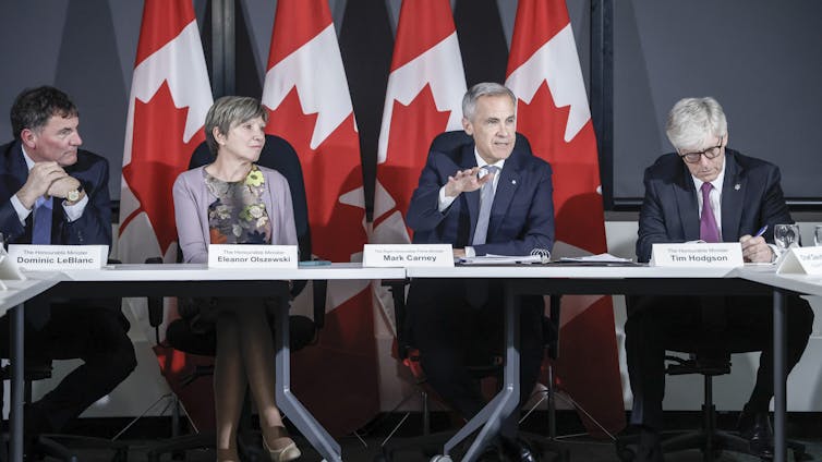 A man with short grey hair sits at a long table with two other men and a woman with a row of Canadian flags behind them.