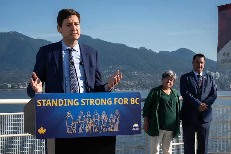 A man in a blue suit speaks at a podium, a woman in a green jacket and another man stand behind him, mountains can be seen in the distance behind them.