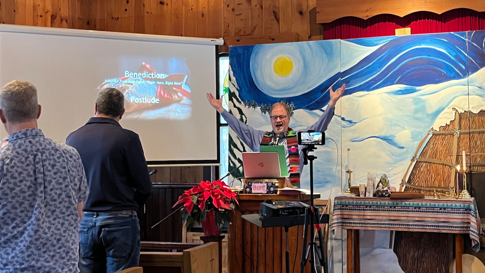 A man with raised arms talks to people in pews in a small wooden church.